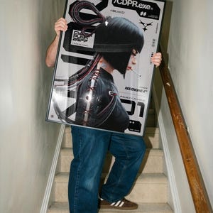 Op de afbeelding: Een ingelijste poster met een futuristische vrouw met zwart haar en een sigaret, tegen een zilveren en zwarte achtergrond. De poster bevat de tekst "/CDPR.exe" en "Cyberpunk 2077". De persoon draagt een blauwe spijkerbroek en bruine sneakers.