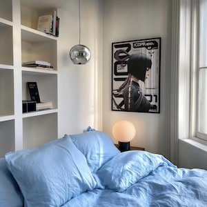 Op de afbeelding: Een slaapkamer met lichtblauw beddengoed, een verchroomde hanglamp en een ingelijste poster met de tekst "/CDPR.exe". Een kleine, ronde lamp staat op een nachtkastje. Witte inbouwkasten bevatten boeken en decoratieve items.