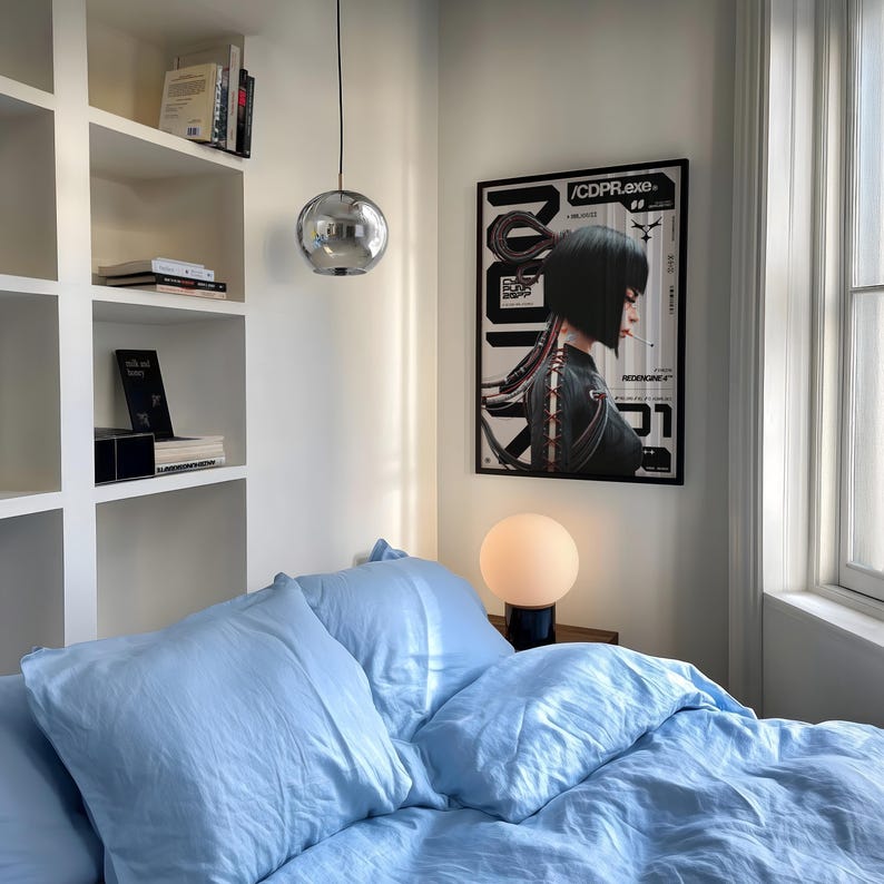 Op de afbeelding: Een slaapkamer met lichtblauw beddengoed, een verchroomde hanglamp en een ingelijste poster met de tekst "/CDPR.exe". Een kleine, ronde lamp staat op een nachtkastje. Witte inbouwkasten bevatten boeken en decoratieve items.