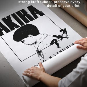 Op de afbeelding: Zwart-wit poster met het woord "AKIRA" in grote letters. De poster wordt opgerold, met de tekst "DESIGNED TO TRAVEL SAFELY" zichtbaar. Het is opgerold en verzegeld in een kraftkoker.