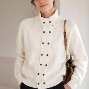 May include: A cream-colored knit sweater featuring a high mock neck collar and a vertical double-breasted button placket down the center front. The sweater has long sleeves with ribbed cuffs and a ribbed hem, accented by dark brown circular buttons.