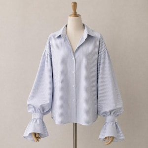 Puede incluir: Camisa a rayas azules y blancas con botones, mangas largas abullonadas y puños acampanados. La camisa se exhibe en un maniquí, mostrando su diseño y ajuste. El cuello es de estilo clásico.