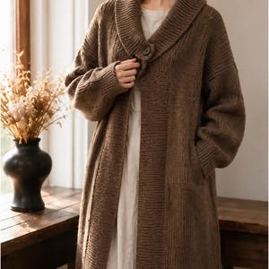May include: A long, brown, open-front knit cardigan featuring a wide shawl collar, long sleeves, and a single button closure at the neckline. The garment is crafted with a ribbed texture and includes deep side pockets, worn over a plain light-colored shirt.