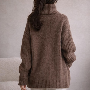 May include: A brown turtleneck sweater with ribbed detailing on the sleeves and body. The sweater is made of a soft, textured material and has a relaxed fit. The model is wearing white pants. The sweater is a warm and cozy garment.