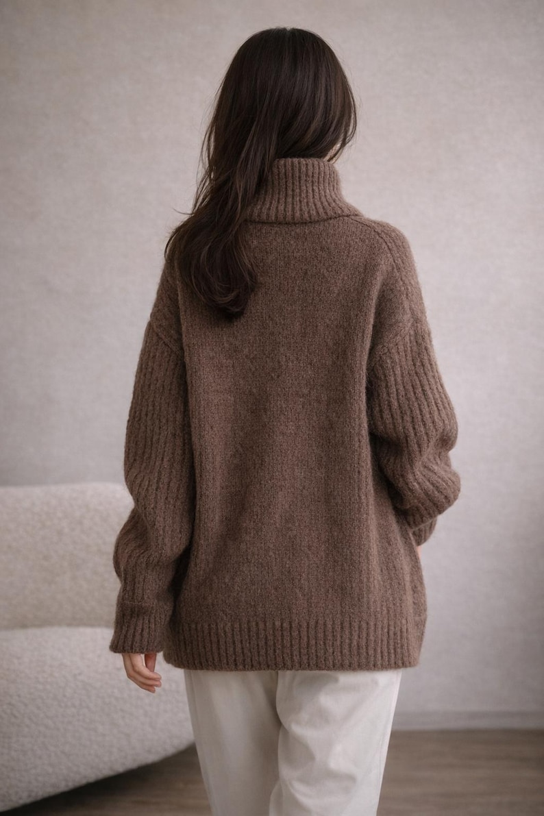 May include: A brown turtleneck sweater with ribbed detailing on the sleeves and body. The sweater is made of a soft, textured material and has a relaxed fit. The model is wearing white pants. The sweater is a warm and cozy garment.