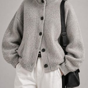 Puede incluir: Cárdigan gris claro con botones negros y cuello alto. El suéter de punto acanalado se combina con pantalones blancos y un bolso negro. El estilo es casual y cómodo, adecuado para el uso diario.