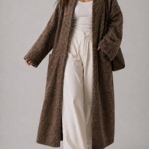 May include: A long, brown, open-front cardigan with two front pockets. The cardigan is worn with a white vest top and cream-coloured wide-leg trousers. A brown shoulder bag completes the outfit. The cardigan appears to be made of a soft, textured material.