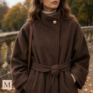 May include: A dark brown wool-blend coat featuring a high collar, a single gold-toned button closure at the neck, and a matching tie belt at the waist. The coat has long sleeves with cuffs and is worn over a cream-colored ribbed turtleneck sweater.