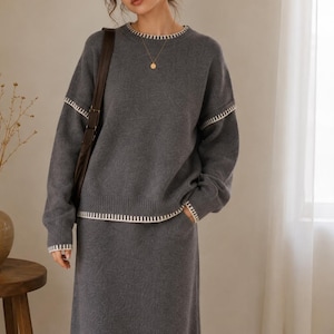 May include: A charcoal gray knit sweater and matching skirt set featuring a crew neckline and long sleeves. The garment is accented with white whipstitch detailing along the collar, cuffs, hemline, and the horizontal seams across the upper sleeves.