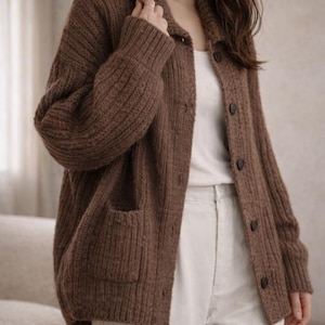 May include: A brown, button-down cardigan sweater with ribbed knit texture. The sweater has a collared neckline, two front pockets, and dark buttons. The model is wearing a white tank top and cream-colored pants.