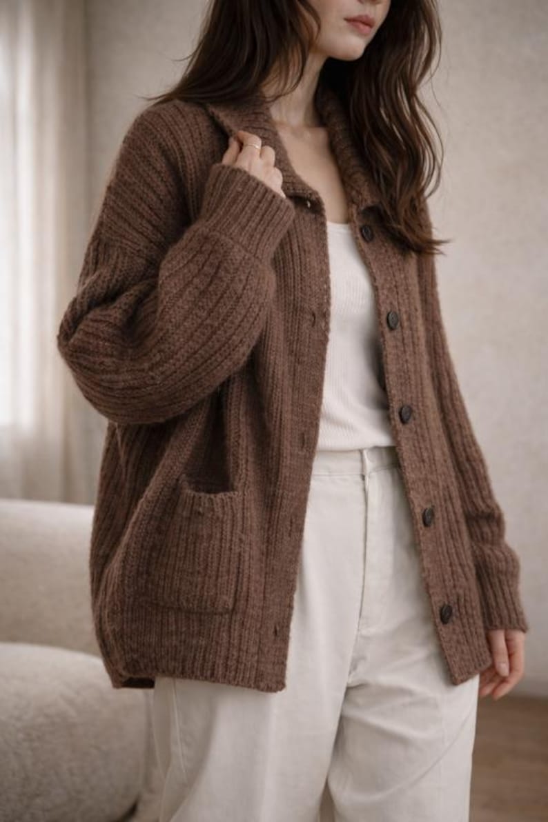 May include: A brown, button-down cardigan sweater with ribbed knit texture. The sweater has a collared neckline, two front pockets, and dark buttons. The model is wearing a white tank top and cream-colored pants.