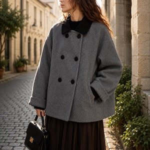 Può includere: Un cappotto grigio doppiopetto con colletto e bottoni neri. Il cappotto ha una vestibilità ampia e maniche con risvolto. Una borsa nera è tenuta in mano. Il cappotto fa parte di una collezione moda autunnale.