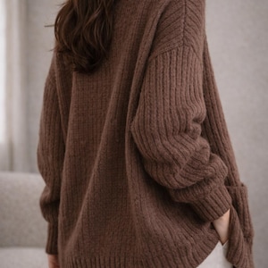 May include: A brown turtleneck sweater with ribbed detailing on the sleeves and body. The sweater has a relaxed fit and is made of a soft, knitted material. The model is wearing white pants.