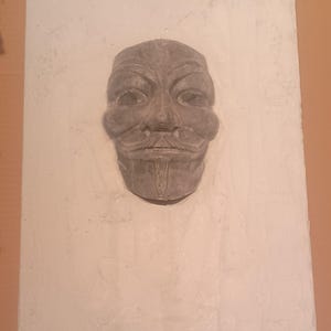 Könnte beinhalten: Eine grau geformte Maske mit stilisiertem Gesicht, mit einem markanten Schnurrbart und Ziegenbart. Die Maske ist vor einem strukturierten, cremefarbenen Hintergrund platziert, was einen auffälligen Kontrast erzeugt. Das Design der Maske erinnert an die Guy-Fawkes-Maske.