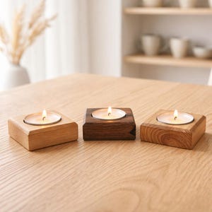 Könnte beinhalten: Drei hölzerne Kerzenhalter mit brennenden Kerzen auf einem hellen Holztisch. Die Kerzenhalter sind quadratisch, einer aus hellem Holz, einer aus dunklem Holz und einer in einem mittleren Farbton. Der Hintergrund umfasst eine Vase mit Trockenblumen und ein Regal mit weißem Geschirr.