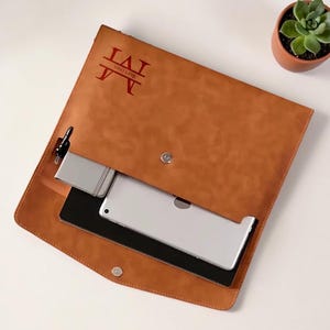 May include: A brown leather portfolio with a personalized red monogram. The open portfolio reveals a tablet, phone, and pen. A small potted succulent sits nearby. The portfolio is designed to hold electronic devices and accessories.