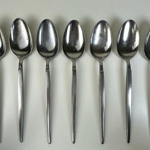 Könnte beinhalten: Sieben Edelstahl-Löffel in einer Reihe angeordnet. Jeder Löffel hat eine ovale Schüssel und einen langen, schlanken Stiel. Die Löffel bestehen aus einem glänzenden, silberfarbenen Metall, das das Licht reflektiert.