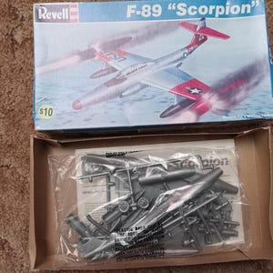 Puede incluir: Caja de maqueta del avión Revell F-89 "Scorpion". La caja muestra un avión a reacción plateado y rojo con el texto "USA AIR FORCE". Contiene una bolsa con piezas de modelo de plástico sin montar.
