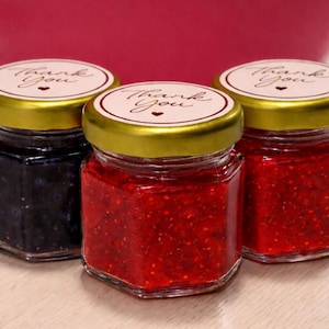 May include: Three small glass jars filled with preserves. The jars have gold lids and labels that read "Thank You." The preserves are red and dark purple. The jars are on a light-colored surface.