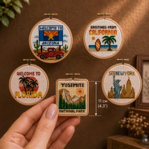 Pode incluir: Cinco bastidores de ponto cruz com temas de viagem: Arizona com um caminhão, Califórnia com uma prancha de surf, Flórida com palmeiras, Nova York com a Estátua da Liberdade e um bastidor quadrado do Yosemite de 11 cm.