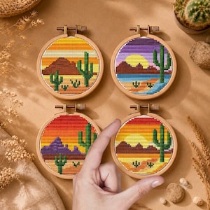 Puede incluir: Cuatro bastidores de bordado circulares de madera con paisajes desérticos en punto de cruz. Cada bastidor presenta una paleta de colores de atardecer única, incluyendo naranja, morado, rojo y amarillo, con cactus saguaro y montañas.