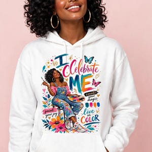 Può includere: Una felpa con cappuccio bianca caratterizzata da una stampa grafica colorata di una persona con capelli ricci seduta su uno sgabello, che tiene in mano un pennello e una tavolozza. Il testo recita I Celebrate ME, create your magic, paint your joy e live in COLOR.