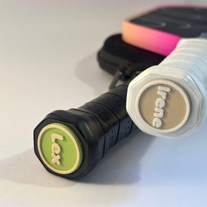 May include: Two pickleball paddles with custom end caps. One paddle has a white grip and a beige end cap with the name Irene. The other paddle has a black grip and a lime green end cap with the name Lex. The paddle faces are black with pink edges.