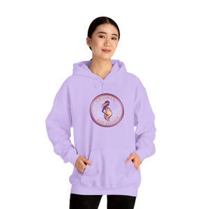May include: A light purple hoodie with a drawstring hood and a kangaroo pocket. The front features a circular graphic with the words "SHOWIN AND GLOWIN" around an illustration of a pregnant person. The hoodie is made of a soft material.