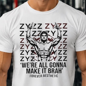 Può includere: Una t-shirt bianca a maniche corte con una stampa grafica di un bodybuilder con le braccia alzate. Lo sfondo della stampa è costituito dal testo ripetuto ZYZZ in nero e bordeaux. Sotto la figura si trova il testo WE'RE ALL GONNA MAKE IT BRAH.