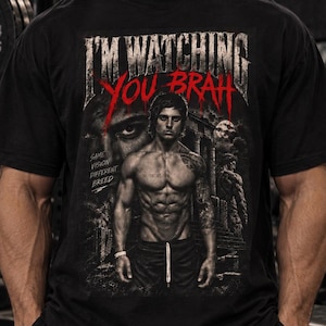 Könnte beinhalten: Ein schwarzes Kurzarm-T-Shirt mit einem Grafikdruck, der den Text I'M WATCHING YOU BRAH in Weiß und Rot zeigt, sowie ein zentrales Bild einer muskulösen Figur und den Text SAME VISION DIFFERENT BREED in einem dunklen, abgenutzten Stil.