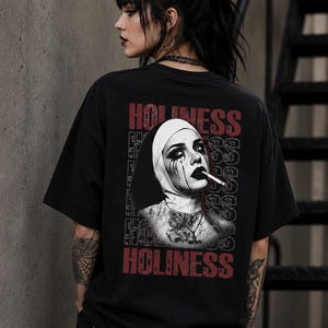 May include: A black short-sleeve t-shirt featuring a graphic print on the back. The design displays a black and white portrait of a nun with dark makeup, smoking a cigarette, surrounded by the repeated red and white text HOLINESS.