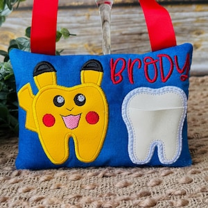 May include: A blue tooth fairy pillow with a yellow Pikachu-shaped tooth design and a white tooth pocket. The name "Brody" is embroidered in red. Red ribbon handles are attached to the top.