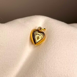 以下が含まれることがあります： 詳細なレイヤーデザインの金色のハート型ペンダント。ペンダントの上部にはチェーン用の小さなループが付いています。ハートは柔らかい白い背景に配置されています。