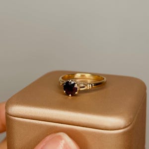 Op de afbeelding: Een gouden ring met een enkele ronde, dieprode edelsteen in een zetting met zes pootjes. De ringband is glad en gepolijst, met een decoratief gespleten detail waar deze de zetting ontmoet, wat een klassiek ontwerp met een donkere steen toont.