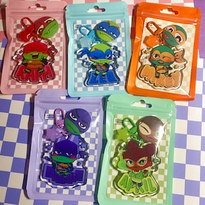 May include: Five acrylic keychains in individual resealable pouches, each featuring a cartoon character from the Teenage Mutant Ninja Turtles franchise. The characters include Raph, Leo, Mikey, Donnie, and April, each with a matching star-shaped clasp.