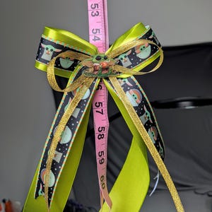 Könnte beinhalten: Eine handgefertigte Haarschleife mit einem grünen und schwarzen Band mit goldenem Besatz und einem Baby Yoda-Charakterdesign. Die Schleife ist mit einem Baby Yoda-Charm akzentuiert. Ein rosa Maßband befindet sich hinter der Schleife, mit Messungen in Zentimetern.