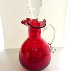 May include: A vibrant red glass cruet with a clear glass handle and stopper. The cruet has a rounded body, a flared rim, and a clear, teardrop-shaped stopper. The handle features a decorative glass bead. This decorative piece is ideal for serving oil or vinegar.