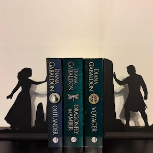 May include: Black metal bookends featuring silhouettes of a woman and a man, framing three books. The books are titled "Outlander", "Dragonfly in Amber", and "Voyager" by Diana Gabaldon. The bookends and books are on a black surface.