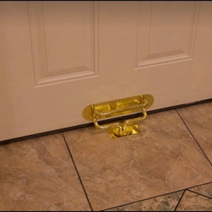 Puede incluir: Un tope de puerta de color latón, montado en el suelo debajo de una puerta blanca. El tope de la puerta tiene un asa y una base plana. El suelo está embaldosado con un patrón beige y marrón.