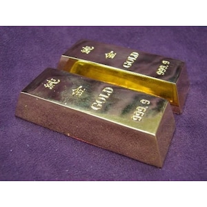 May include: Two gold bars, each with the word "GOLD" and "999.9" stamped on them. The bars are rectangular and have Chinese characters on them. The bars are on a purple surface.