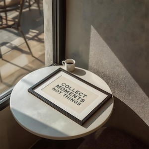 May include: A rectangular black picture frame containing a cream-colored print with the black text COLLECT MOMENTS NOT THINGS. The frame rests on a round white table next to a small white ceramic cup filled with a dark liquid beverage.
