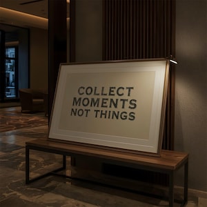May include: A rectangular framed wall art piece featuring a light beige background with the dark gray text COLLECT MOMENTS NOT THINGS centered in a bold, sans-serif font. The artwork is enclosed in a thin, metallic gold frame with a white mat border.