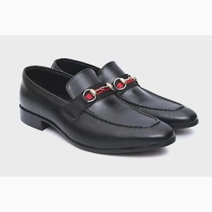 May include: A pair of black leather loafers featuring a prominent strap across the vamp. The strap is accented with a horizontal red stripe and a gold-toned metal horsebit detail. The shoes have a smooth finish, a rounded toe, and a low heel.