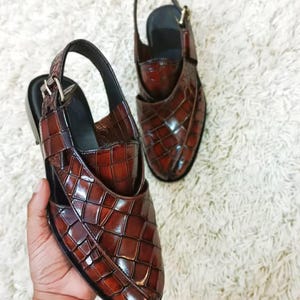 Luxury brown crocodile-textured leather slingback sandals for men with pointed toe and buckle strap detail, handmade premium formal footwear styled with beige trousers in an elegant indoor setting.