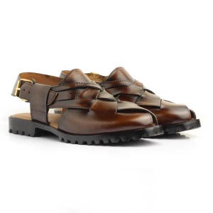 May include: A pair of brown leather sandals with a woven design. The sandals have a thick, black sole and a gold-colored buckle. The leather has a glossy finish. The sandals are displayed on a white background.