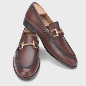 Puede incluir: Un par de mocasines de cuero marr&oacute;n con detalles met&aacute;licos dorados. Los zapatos tienen un dise&ntilde;o cl&aacute;sico sin cordones con una hebilla decorativa en la pala. El cuero parece pulido y los zapatos est&aacute;n sobre un fondo neutro.