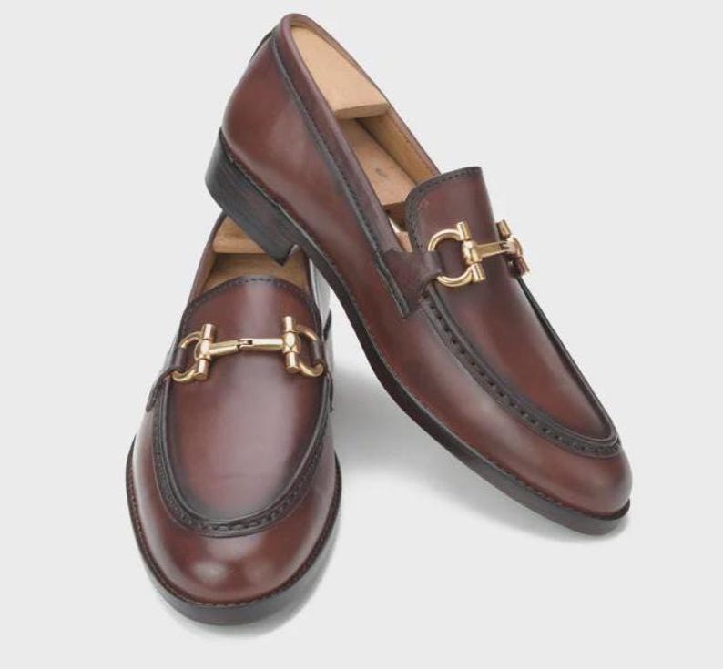 Puede incluir: Un par de mocasines de cuero marr&oacute;n con detalles met&aacute;licos dorados. Los zapatos tienen un dise&ntilde;o cl&aacute;sico sin cordones con una hebilla decorativa en la pala. El cuero parece pulido y los zapatos est&aacute;n sobre un fondo neutro.