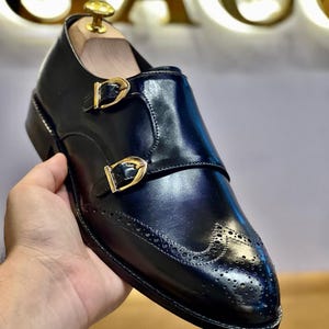 May include: Black leather double monk strap shoe with gold-coloured buckles. The shoe has decorative perforations on the toe and a polished finish. A wooden shoe tree is inside the shoe.
