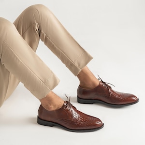 Könnte beinhalten: Braune Lederschuhe mit einem strukturierten Muster auf der Spitze und dem Vorderblatt. Die Schuhe haben ein klassisches Schnürdesign und eine schwarze Sohle. Das Bild zeigt die Schuhe von unten, mit den Beinen des Trägers in beigefarbenen Hosen.