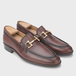 Puede incluir: Un par de mocasines de cuero marr&oacute;n con detalles met&aacute;licos dorados. Los zapatos tienen un dise&ntilde;o cl&aacute;sico con punta redondeada y tac&oacute;n bajo. Los mocasines se muestran sobre un fondo neutro.